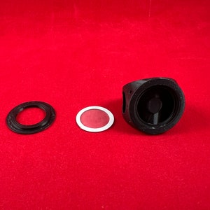 May include: Three black plastic parts for a 3D printed camera lens. The parts include a round ring, a flat disc with a red centre, and a larger round piece with a circular opening.