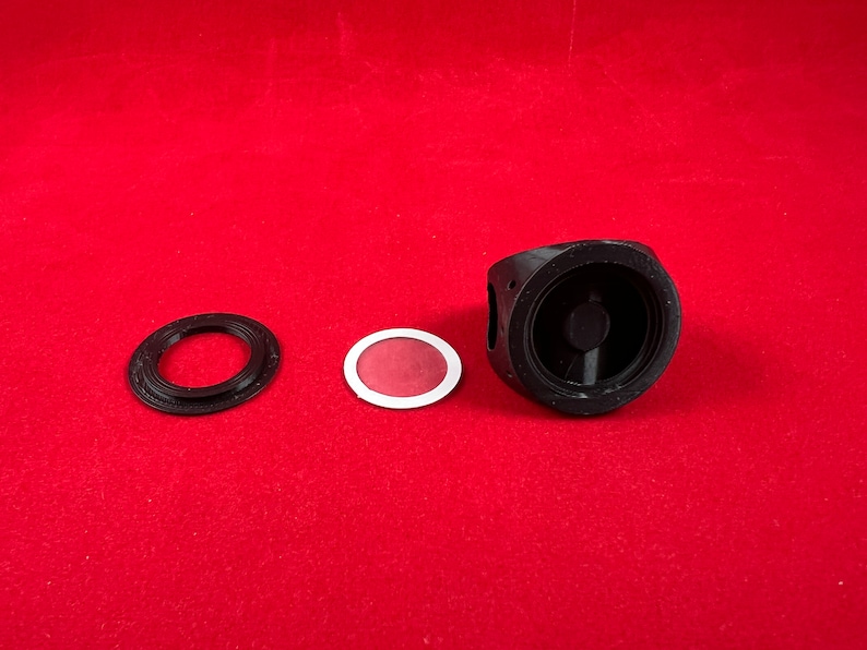 May include: Three black plastic parts for a 3D printed camera lens. The parts include a round ring, a flat disc with a red centre, and a larger round piece with a circular opening.