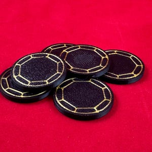 May include: A stack of six black poker chips with gold trim. The chips have a textured surface and a geometric design.