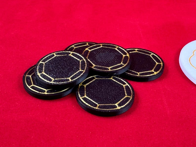 May include: A stack of six black poker chips with gold trim. The chips have a textured surface and a geometric design.