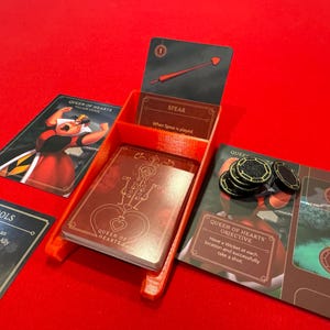 May include: A red plastic game organizer with a Queen of Hearts card, a Spear card, and a Queen of Hearts objective card. The organizer has a slot for each card and a space for tokens.