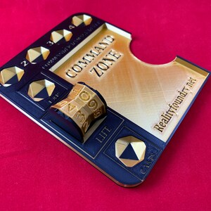 May include: A black and gold 3D printed game board with a command zone, life, and cast area. The board has a gold die with numbers 1-9 and a space for a card. The board is labeled "Realityfoundry.net".