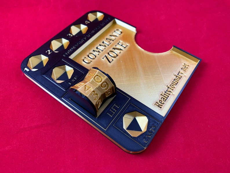 May include: A black and gold 3D printed game board with a command zone, life, and cast area. The board has a gold die with numbers 1-9 and a space for a card. The board is labeled "Realityfoundry.net".