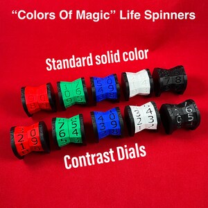 May include: Eight life counters, or spinners, in various colors, including red, blue, green, white, and black. Each counter has a number dial with numbers 0-9. The counters are labeled "Standard solid color" and "Contrast Dials".