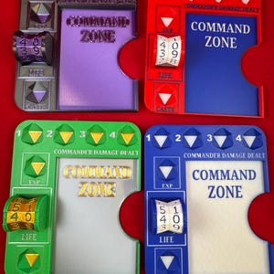 May include: Four rectangular game tracking boards in purple, red, green, and blue. Each board features a central Command Zone area, a rotating life counter dial, and numbered slots for tracking commander damage, experience, and cast counts.