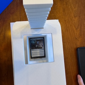 May include: A white foam display stand with a card game card inside. The card is a blue and white Magic: The Gathering card with the text "Chancellor of the Spires".