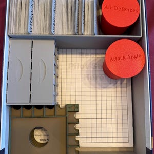 May include: A game box organizer with compartments for cards, tokens, and a gridded game board. The organizer has a gray and brown color scheme. The compartments are labeled "Veteranance", "Average Skill", "Small Size", "Air Defences", and "Attack Angle".