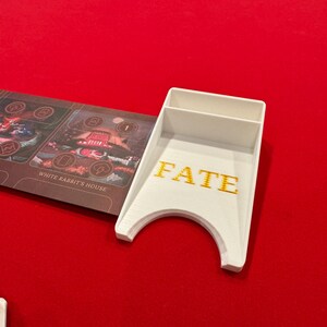 May include: White plastic card holder with the word "FATE" printed in gold letters. The holder has a slot for cards and a curved bottom to hold dice.