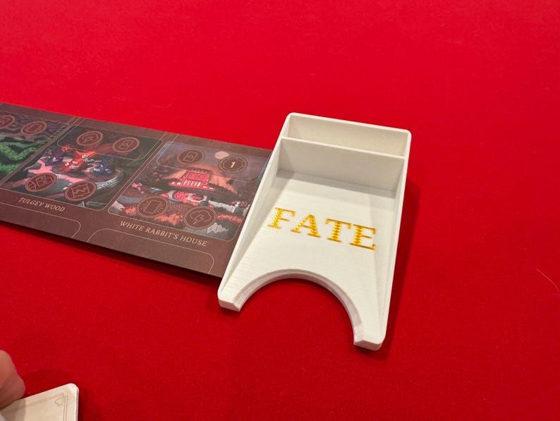 May include: White plastic card holder with the word "FATE" printed in gold letters. The holder has a slot for cards and a curved bottom to hold dice.