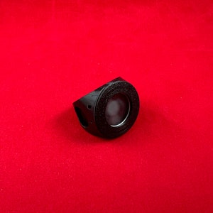 May include: A black plastic ring with a circular opening in the centre. The ring has a textured surface and a small hole on the side.