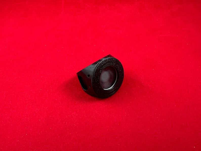 May include: A black plastic ring with a circular opening in the centre. The ring has a textured surface and a small hole on the side.