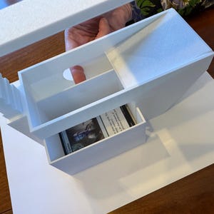May include: White foam board model of a building with a card inside a drawer. The building has a staircase and a small opening in the top.