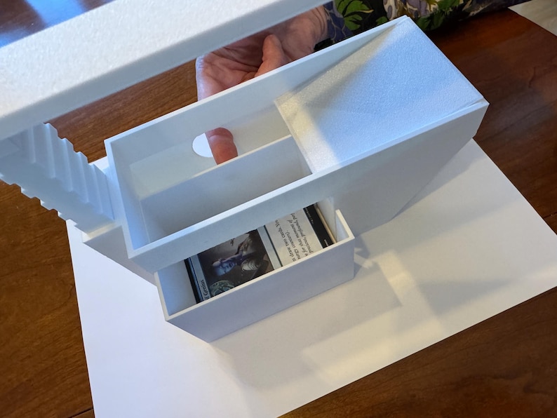 May include: White foam board model of a building with a card inside a drawer. The building has a staircase and a small opening in the top.