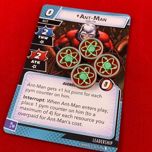 Op de afbeelding: Een blauwe en witte kaart uit een kaartspel met Ant-Man, een superheld in een rood-zwart pak. De kaart heeft een kracht van 0, een sterkte van 2 en een snelheid van 2. De tekst op de kaart luidt: "Ant-Man krijgt +1 trefferpunt voor elke Pym-teller op hem. Onderbreken: Wanneer Ant-Man in het spel komt, plaats 1 Pym-teller op hem (tot maximaal 4) voor elke bron die je te veel hebt betaald voor de kosten van Ant-Man."