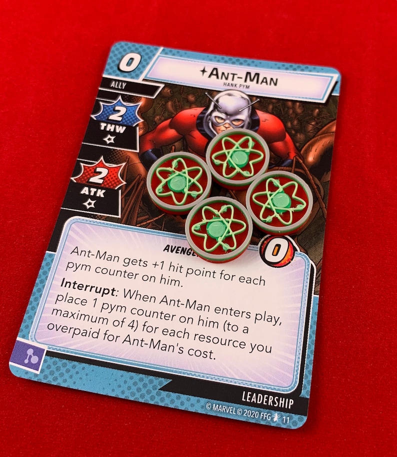 Op de afbeelding: Een blauwe en witte kaart uit een kaartspel met Ant-Man, een superheld in een rood-zwart pak. De kaart heeft een kracht van 0, een sterkte van 2 en een snelheid van 2. De tekst op de kaart luidt: "Ant-Man krijgt +1 trefferpunt voor elke Pym-teller op hem. Onderbreken: Wanneer Ant-Man in het spel komt, plaats 1 Pym-teller op hem (tot maximaal 4) voor elke bron die je te veel hebt betaald voor de kosten van Ant-Man."