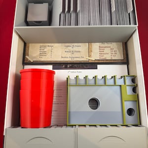Puede incluir: Una caja blanca con un inserto gris para guardar las piezas del juego. La caja contiene un juego de cartas, un juego de contadores y un juego de reglas. La caja está etiquetada con el nombre del juego y el número de edición del juego. La caja también está etiquetada con las reglas del juego y el sistema de puntos del juego.