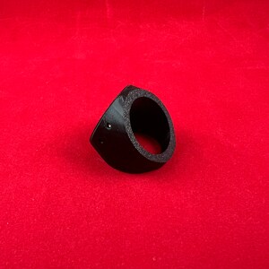 May include: A black plastic ring with a circular opening, designed to hold a speaker or other round object. The ring has a slight angle to it, and there are two small holes on the side.