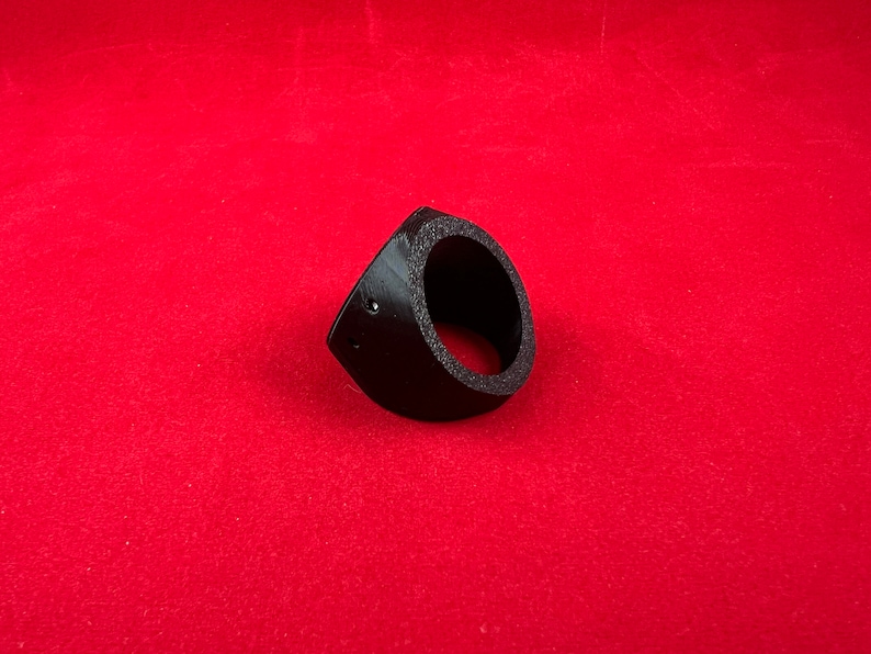 May include: A black plastic ring with a circular opening, designed to hold a speaker or other round object. The ring has a slight angle to it, and there are two small holes on the side.