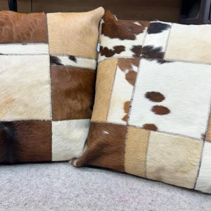 Könnte beinhalten: Zwei quadratische Kissen mit einem Patchwork-Design aus verschiedenen Brauntönen, Weiß und Beige aus Kuhfell.