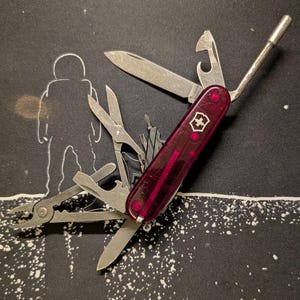 Könnte beinhalten: Ein rotes Multitool mit transparentem Griff und verschiedenen Edelstahlwerkzeugen, darunter ein Messer, eine Schere und ein Flaschenöffner. Das Werkzeug liegt auf einem schwarzen Hintergrund mit einer weißen Illustration eines Astronauten und eines Mondes.