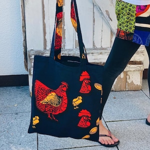 Könnte beinhalten: Eine schwarze Tragetasche mit einem roten und gelben Hühnerdruck. Die Tasche hat einen langen Riemen und wird von einer Person gehalten.