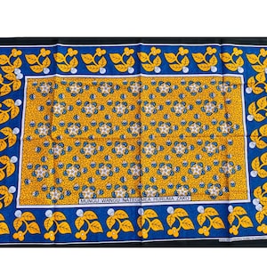 Puede incluir: Un tejido rectangular con un panel central amarillo mostaza con un patrón floral repetitivo en blanco y azul. Está bordeado por un marco azul con hojas amarillas y brotes blancos, e incluye el texto MUNGU WANGU NATEGEMEA HURUMA ZAKO.