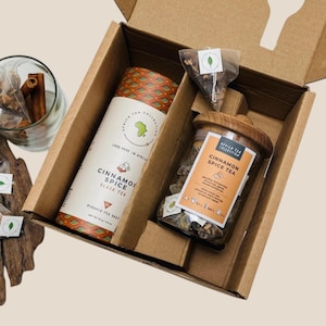 May include: A brown cardboard box with a lid open, revealing a gift set of cinnamon spice tea. The set includes a glass jar of loose leaf tea, a tube of pyramid tea bags, and a small bag of tea. The box is labeled "Africa Tea Collection" and "Cinnamon Spice Tea".