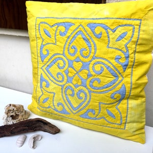May include: A yellow cushion with a blue embroidered design. The cushion is square shaped and made from a soft fabric.