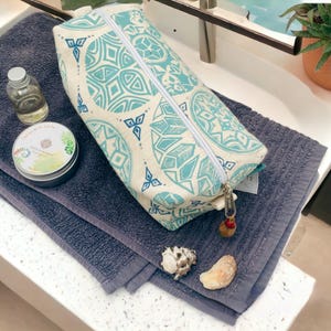 May include: A white wash bag with a blue and white geometric pattern. The bag has a zipper closure and a tassel pull. The bag is sitting on a grey towel.