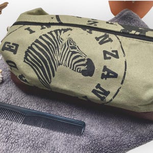 Pode incluir: Uma bolsa de viagem de lona verde com acabamento em couro marrom e fecho de zíper. A bolsa tem uma estampa de zebra em preto e branco e o texto "Tanzânia" impresso nela.