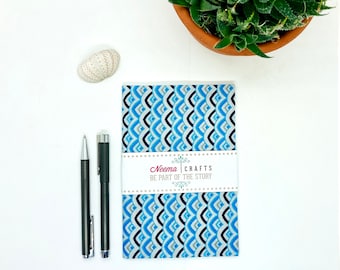 Puede incluir: Un cuaderno con estampado azul y blanco con una etiqueta que dice "Neema Crafts Be Part of the Story". El cuaderno está sobre una superficie blanca con dos bolígrafos y una concha.