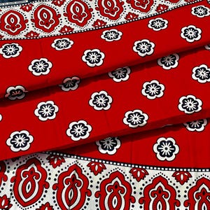 May include: Red fabric with a white and black floral pattern. The fabric features a repeating pattern of white flowers with black centers on a red background. The border has a white and red ornate design. The fabric is folded.