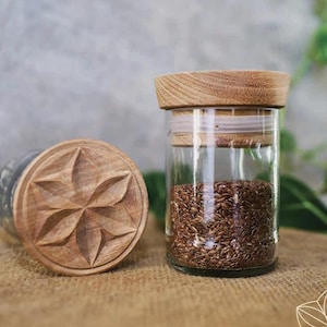 Könnte beinhalten: Zwei zylindrische Vorratsgläser aus klarem Glas mit hellbraunen Holzdeckeln. Ein Deckel liegt flach und zeigt ein geschnitztes, stern- oder blumenförmiges Muster mit sechs Spitzen. Das andere Glas steht aufrecht und ist mit kleinen braunen Samen gefüllt.
