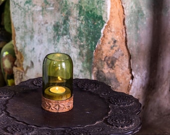May include: A green glass candle holder with a wooden base sits on a dark wooden table with intricate carvings. The candle holder is lit with a small, white candle inside.