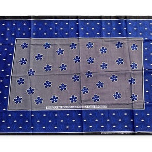 Op de afbeelding: Een blauw en grijs patroontextiel met een bloemenontwerp. Het textiel heeft een donkerblauwe rand met witte stippen en een grijs middenstuk met blauwe bloemmotieven. De tekst "MUNGU NI NGUZO HUONGOZA KWA UPENDO" is zichtbaar.