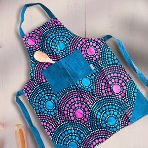 May include: A blue and black apron with a pink and blue circle pattern. The apron has a large blue pocket on the front and a wooden spoon is tucked into the pocket.