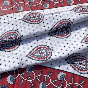 May include: Folded fabric with a white background featuring a repeating pattern of red, blue, and gray paisley-like designs. The edges of the fabric have a red border with floral and leaf patterns.