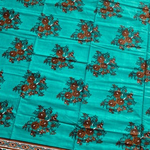 Puede incluir: Una tela turquesa con un estampado floral repetido en marrón y negro. La tela tiene un borde decorativo con un diseño marrón y blanco. El estampado floral está espaciado uniformemente por toda la tela.