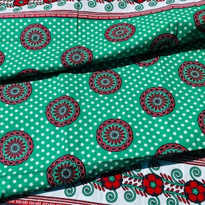 Puede incluir: Una tela doblada con un fondo verde vibrante, un patrón de lunares blancos y motivos circulares repetidos en rojo y verde oscuro. Los bordes están rematados con una franja blanca decorada con flores rojas y enredaderas verdes.
