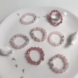 Könnte beinhalten: Sieben rosafarbene und roségoldene Perlenringe mit silbernen Akzenten. Die Ringe sind auf einem weißen Teller mit kleinen weißen Vogelfiguren angeordnet.