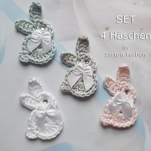 Könnte beinhalten: Set mit vier gehäkelten Hasen-Anhängern in Pastellfarben mit weißen Schleifen. Der Text "SET 4 Häschen in zarten Farben" ist im Bild sichtbar.