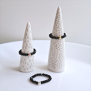 Könnte beinhalten: Drei schwarze Perlenringe mit einem goldenen Perlenakzent auf einem weißen Keramik-Ringständer. Die Ringständer sind kegelförmig mit einem gepunkteten oder gestreiften Muster.