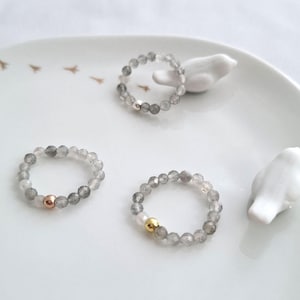 Könnte beinhalten: Drei silberne Ringe mit grauen Edelsteinperlen und einem goldenen Perlenakzent. Die Ringe liegen auf einer weißen Oberfläche.