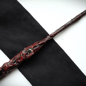 Puede incluir: Una varita artesanal con un acabado texturizado en rojo oscuro y marrón. La varita presenta un detalle central de color plateado y una punta transparente. La varita está sobre una tela negra.