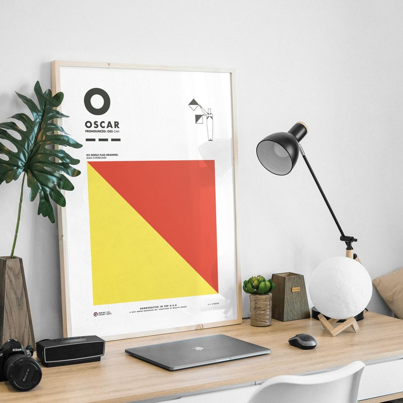 May include: A framed poster with a red and yellow diagonal design. The poster has the word "OSCAR" in black text and the words "PRONOUNCED: OSS-CAR" in smaller black text. The poster also includes a small black and white illustration of a flag.