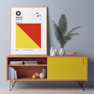 May include: A framed print with a yellow and red geometric design. The print has the word "OSCAR" in black text and the phonetic spelling "pronounced "OS-car" in smaller black text. The print is hanging on a wall above a wooden cabinet with a yellow door.