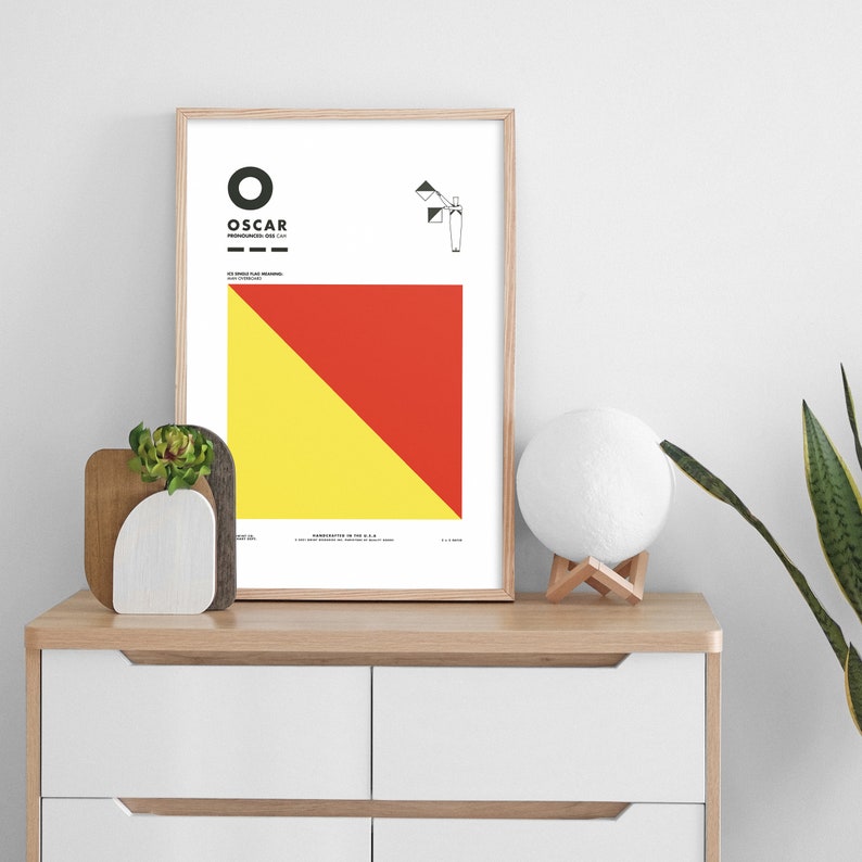 May include: A framed print of the International Maritime Signal Flag "Oscar" with a yellow and red diagonal stripe design. The flag is displayed on a white background with the letter "O" and the word "OSCAR" in black text.