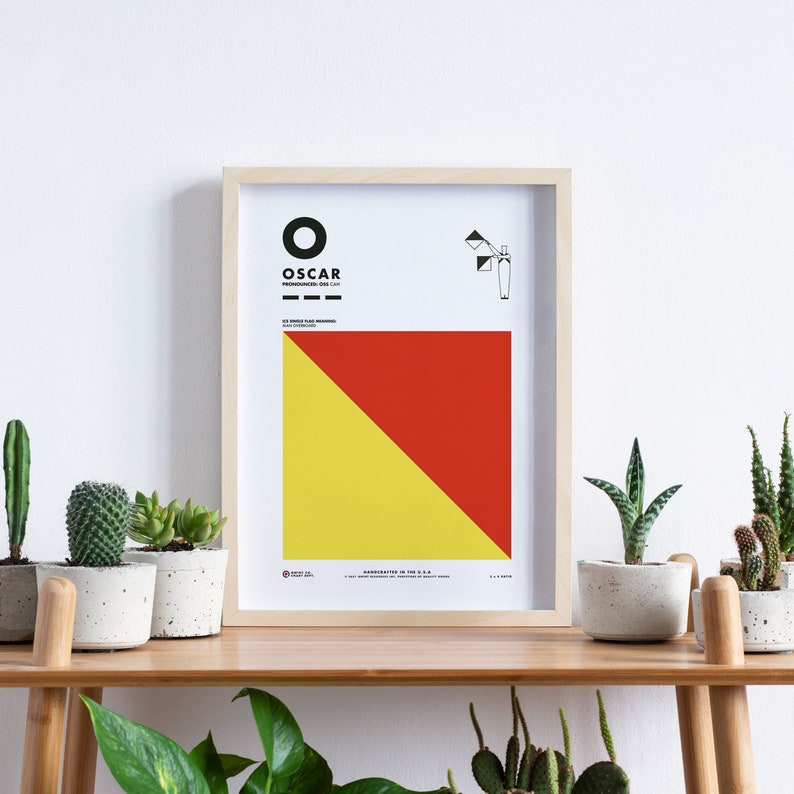 May include: A framed nautical flag chart with the letter "O" and the word "OSCAR" printed in black. The flag is red and yellow with a diagonal line. The chart is framed in natural wood.