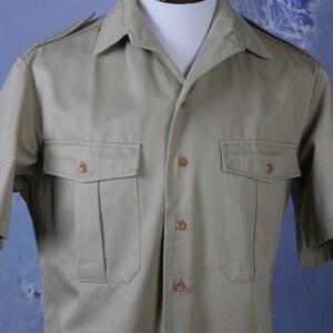 May include: A short-sleeved, light brown utility shirt with a button-down collar and two large flap pockets. The shirt has a button closure and is made of a lightweight fabric.