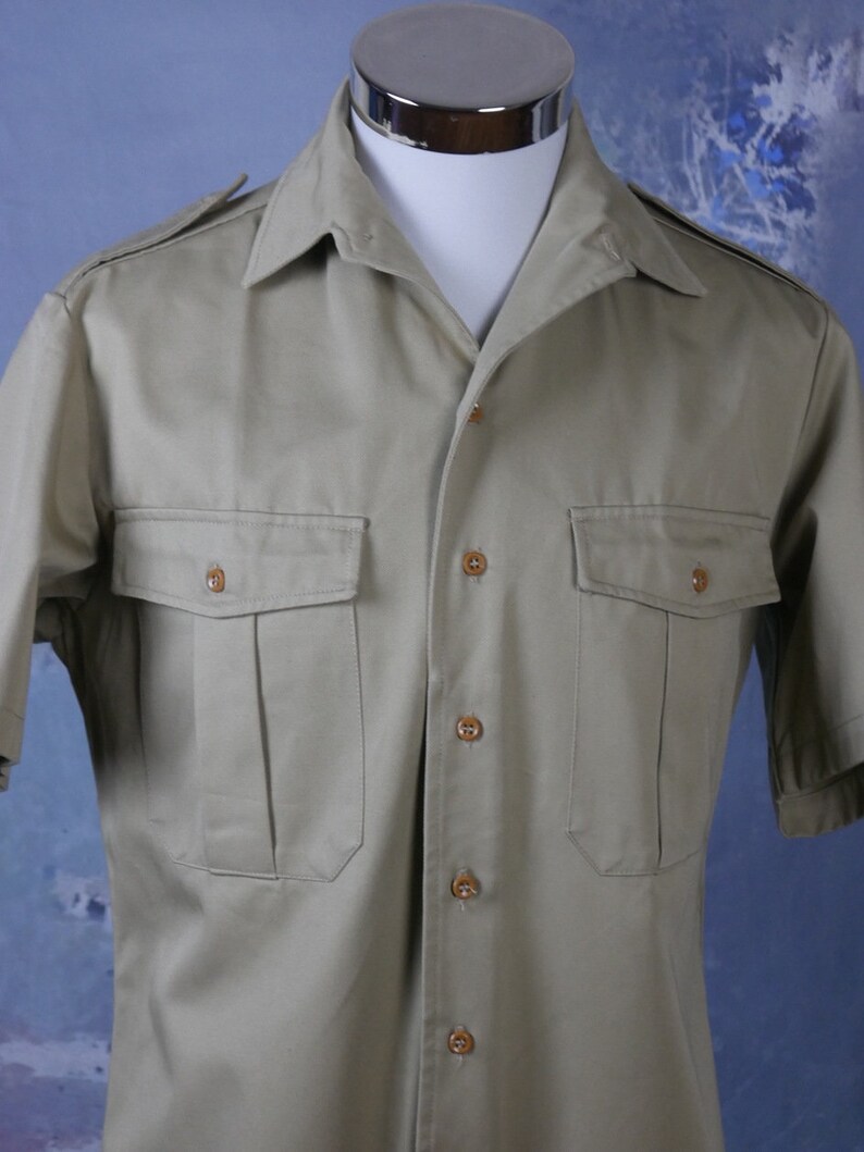 May include: A short-sleeved, light brown utility shirt with a button-down collar and two large flap pockets. The shirt has a button closure and is made of a lightweight fabric.